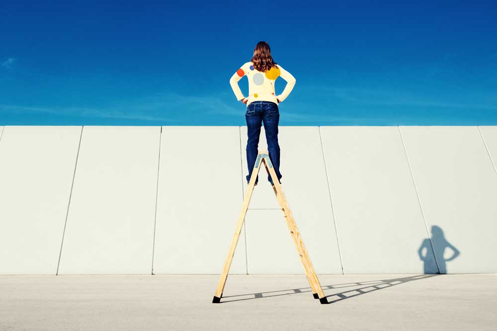 Brand values: curiosity. woman on ladder looking over high fence. blue sky above, concrete fence, concrete foreground
